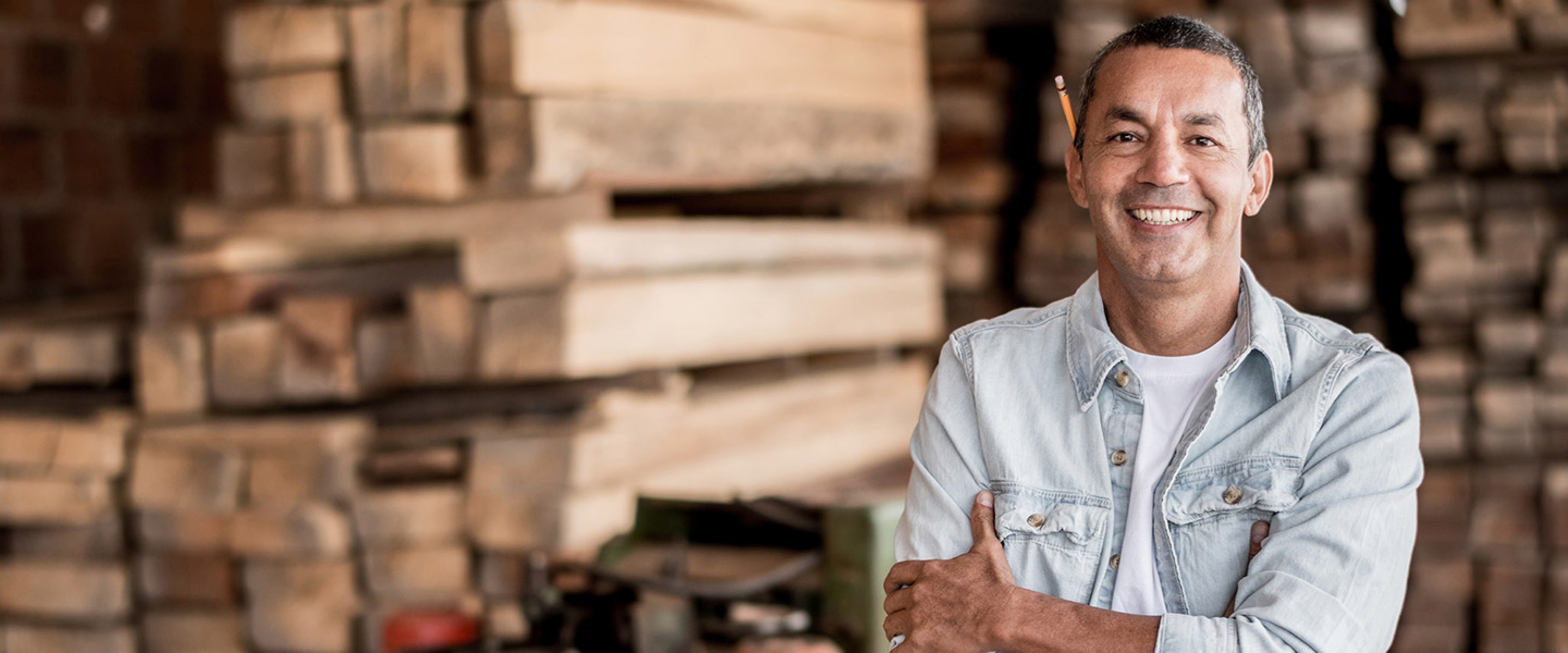 Man with crossed arms and pencil behind his ear smiles in a lumberyard