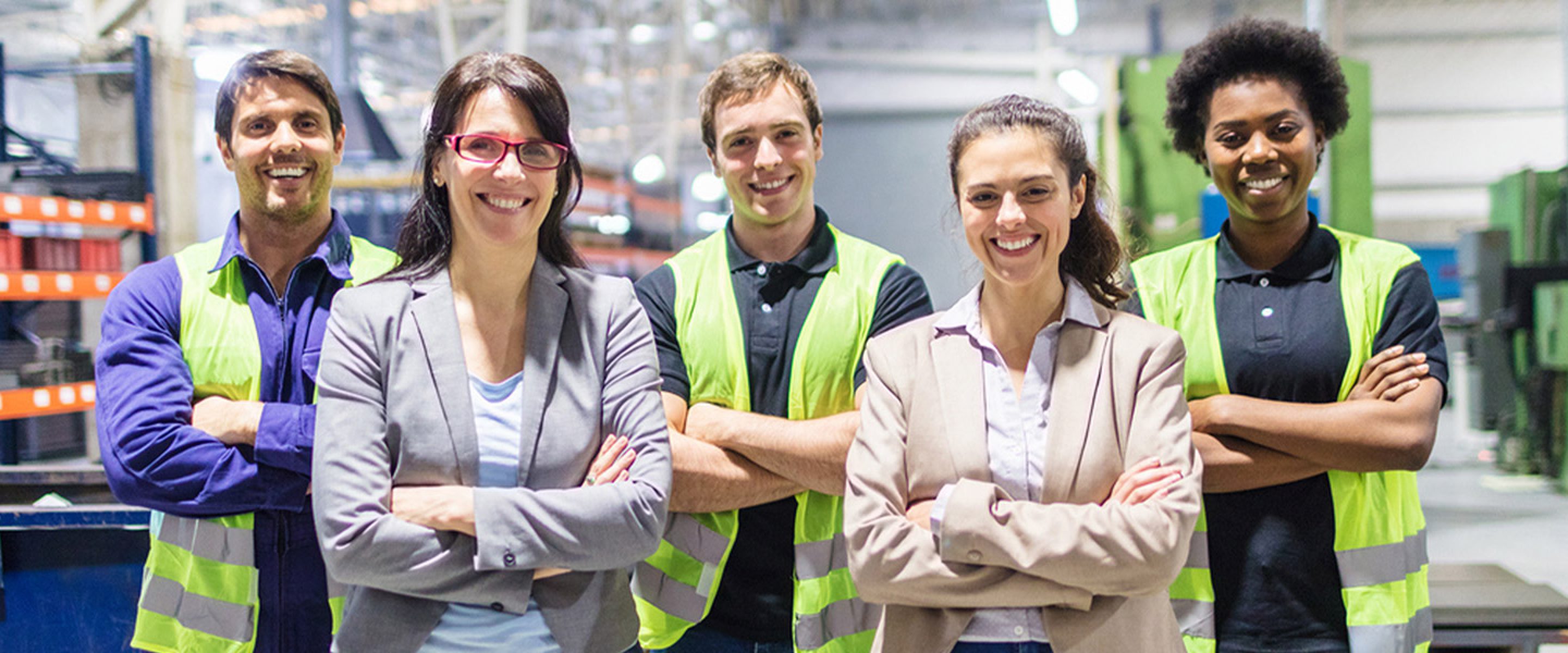 Two business women and three warehouse workers wearing safety vests smile in factory warehouse