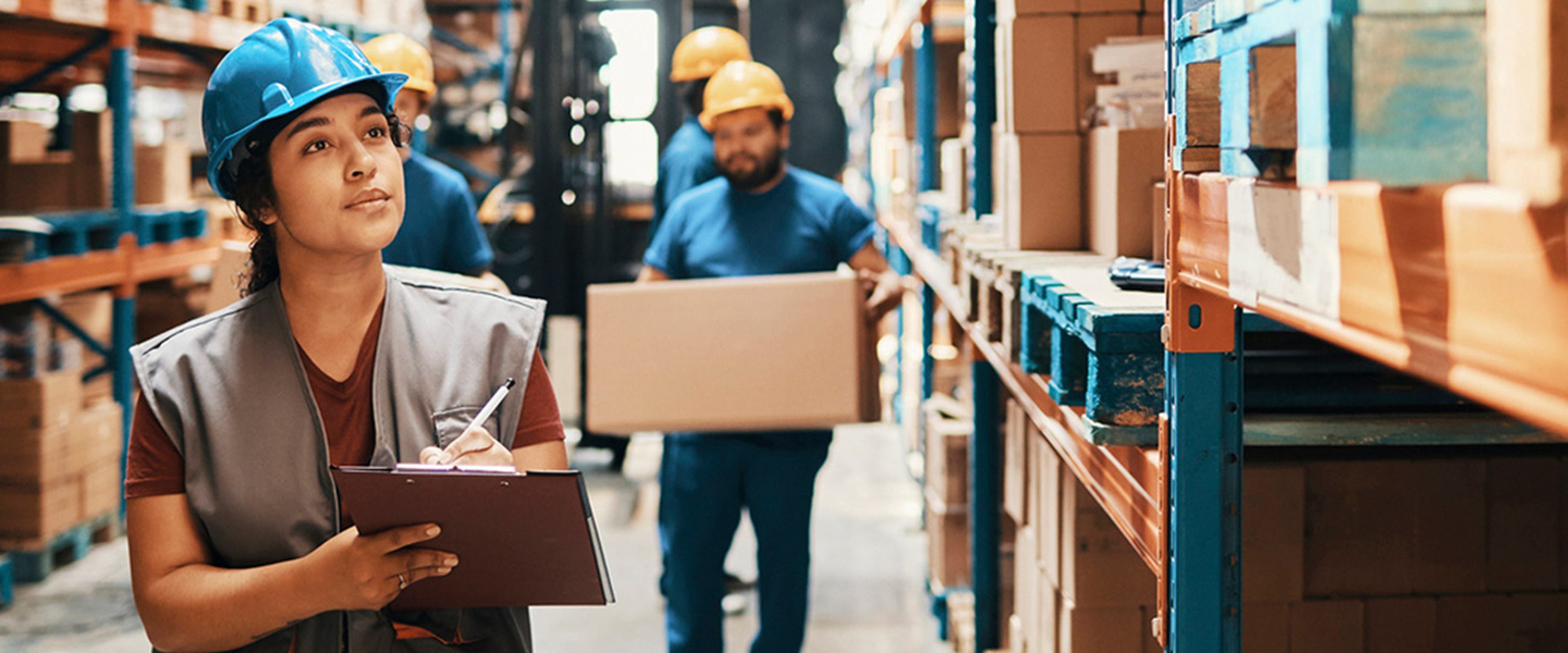 Female warehouse worker writes on her clipboard while other workers carry boxes in the background