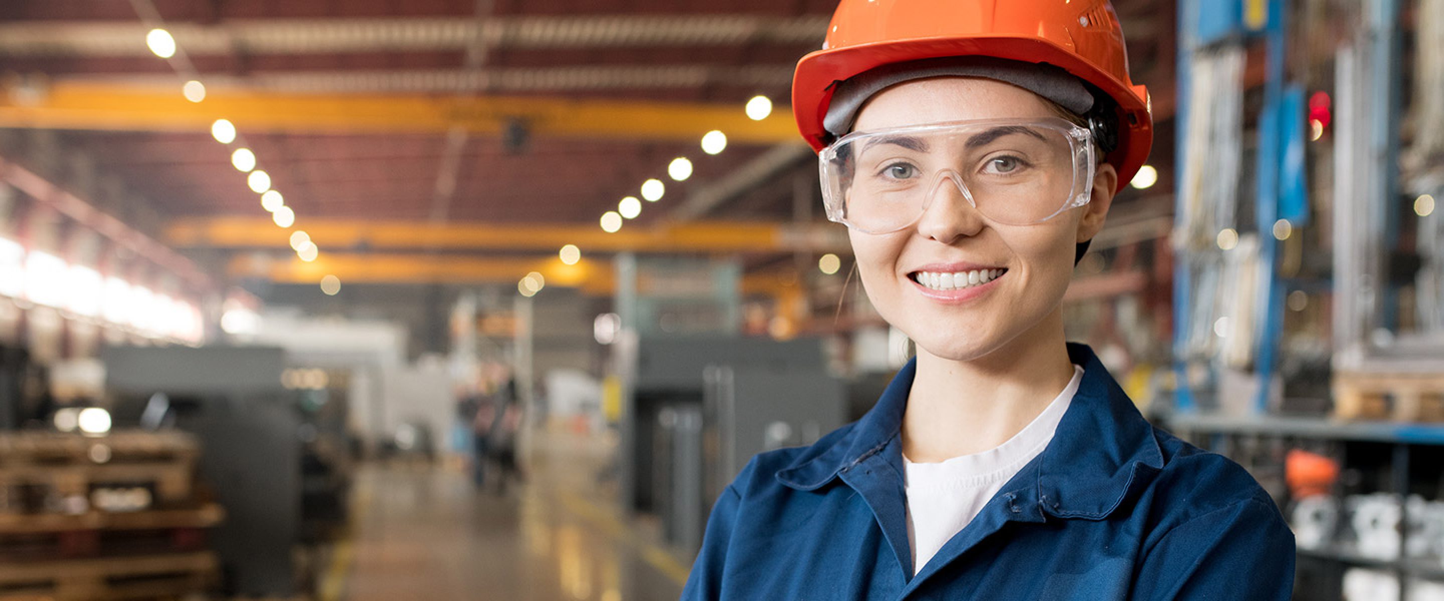 Young female worker wearing safety glasses and a hard hat smiles in a factory